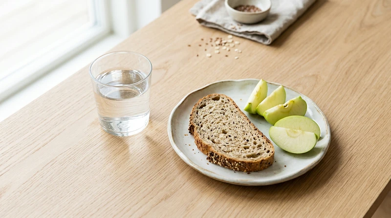 An overhead still life of a glass of water, a piece of bread, and an apple slice on a wooden table