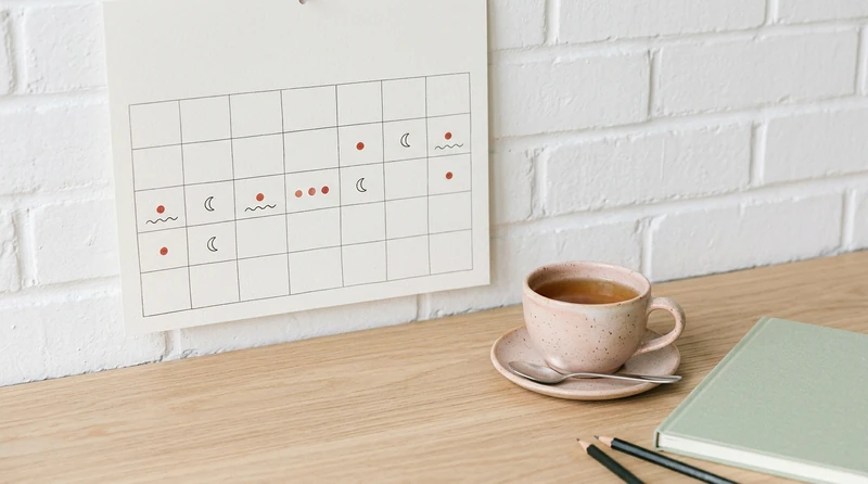 A clean wall calendar marked with small symbols and a cup of tea beside it on a desk